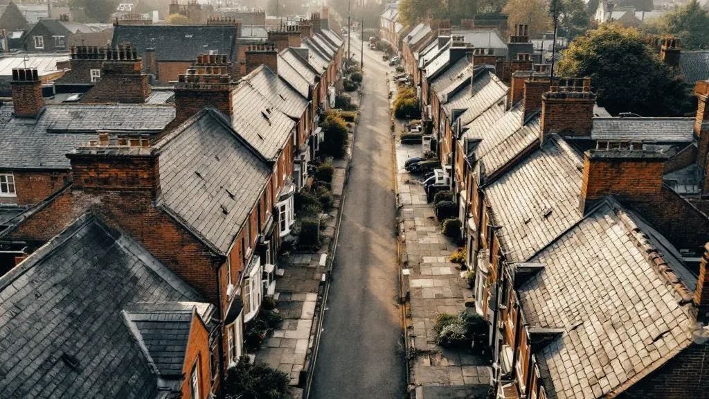 Aerial view of a quiet Victorian terraced street at dawn, lined with uniform brick houses casting long shadows under overcast light, conveying stillness and scarcity.