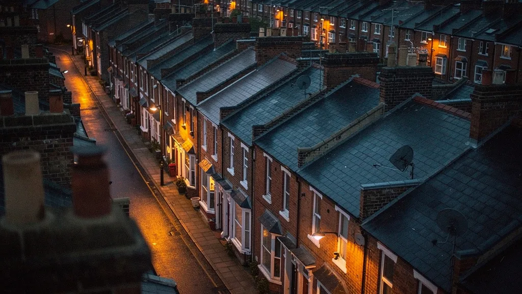 Aerial view at dusk of densely packed Victorian terraced houses with amber streetlights casting long shadows across grey slate rooftops and red-brick facades.