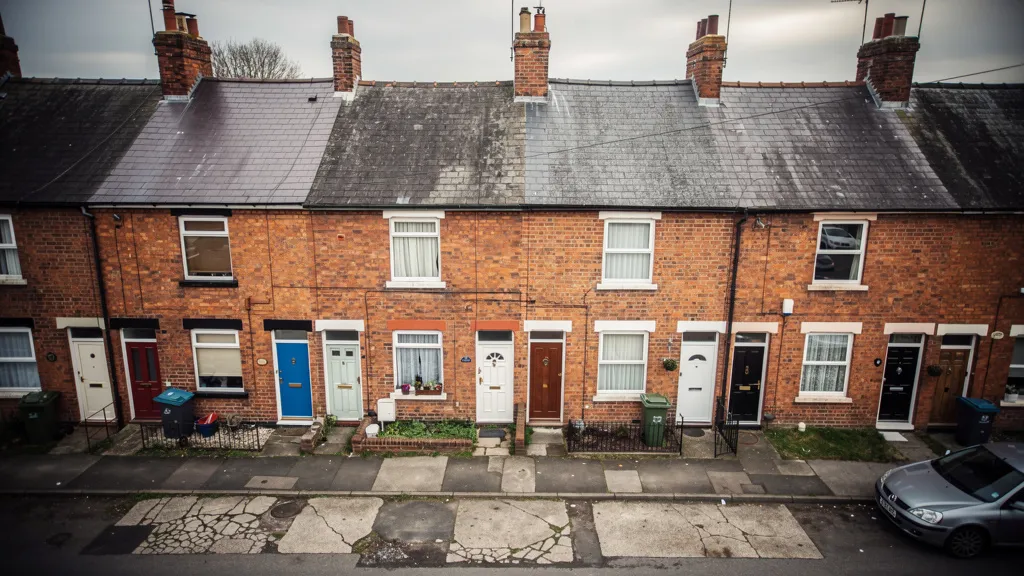 Aerial view of a row of traditional UK terraced houses with red-brick facades and multiple front doors, shot in soft overcast daylight with muted colour tones.