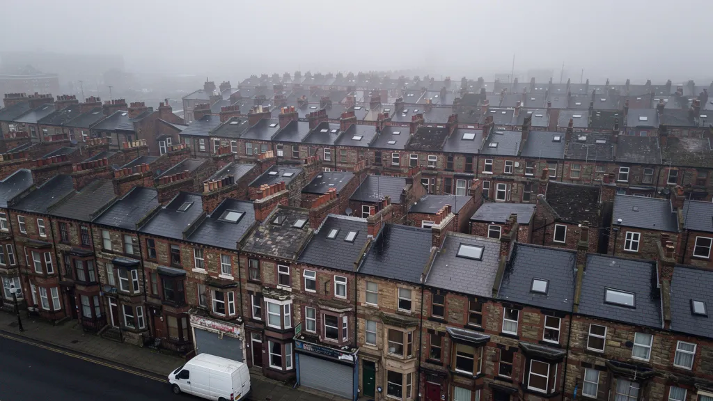 A wide aerial view of rows of traditional British terraced houses in a northern English city, overcast sky, muted brick tones, slight fog in the distance, sense of scale and density, no people visible, photorealistic documentary style