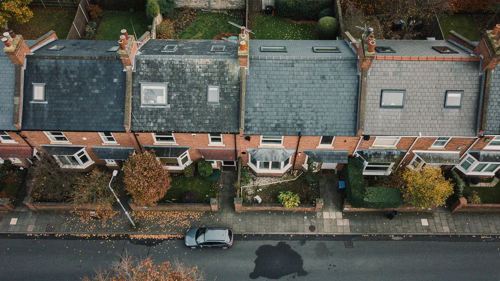 Aerial view of a row of traditional UK terraced houses in a residential street, overcast soft grey sky, subtle autumn colours in small front gardens, muted palette of red brick and grey slate rooftops, quiet and slightly melancholy mood, photorealistic