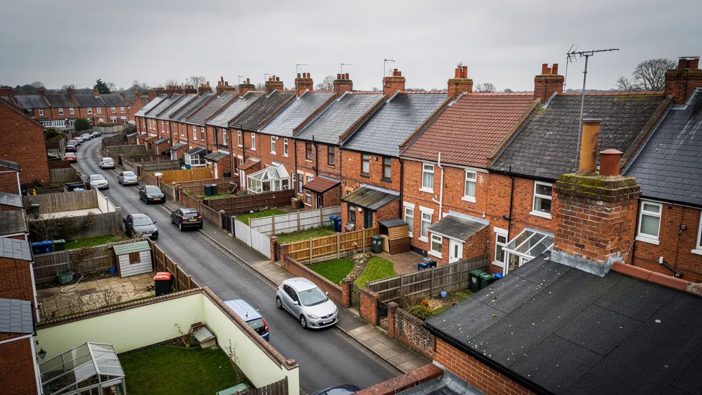 A wide aerial photograph of a row of traditional British terraced houses in a quiet East Midlands town. Red brick rooftops, small back gardens, narrow streets. Overcast sky with soft diffused light. Photorealistic, documentary style, muted warm tones of brick red, slate grey and pale green. No signage, no people visible.